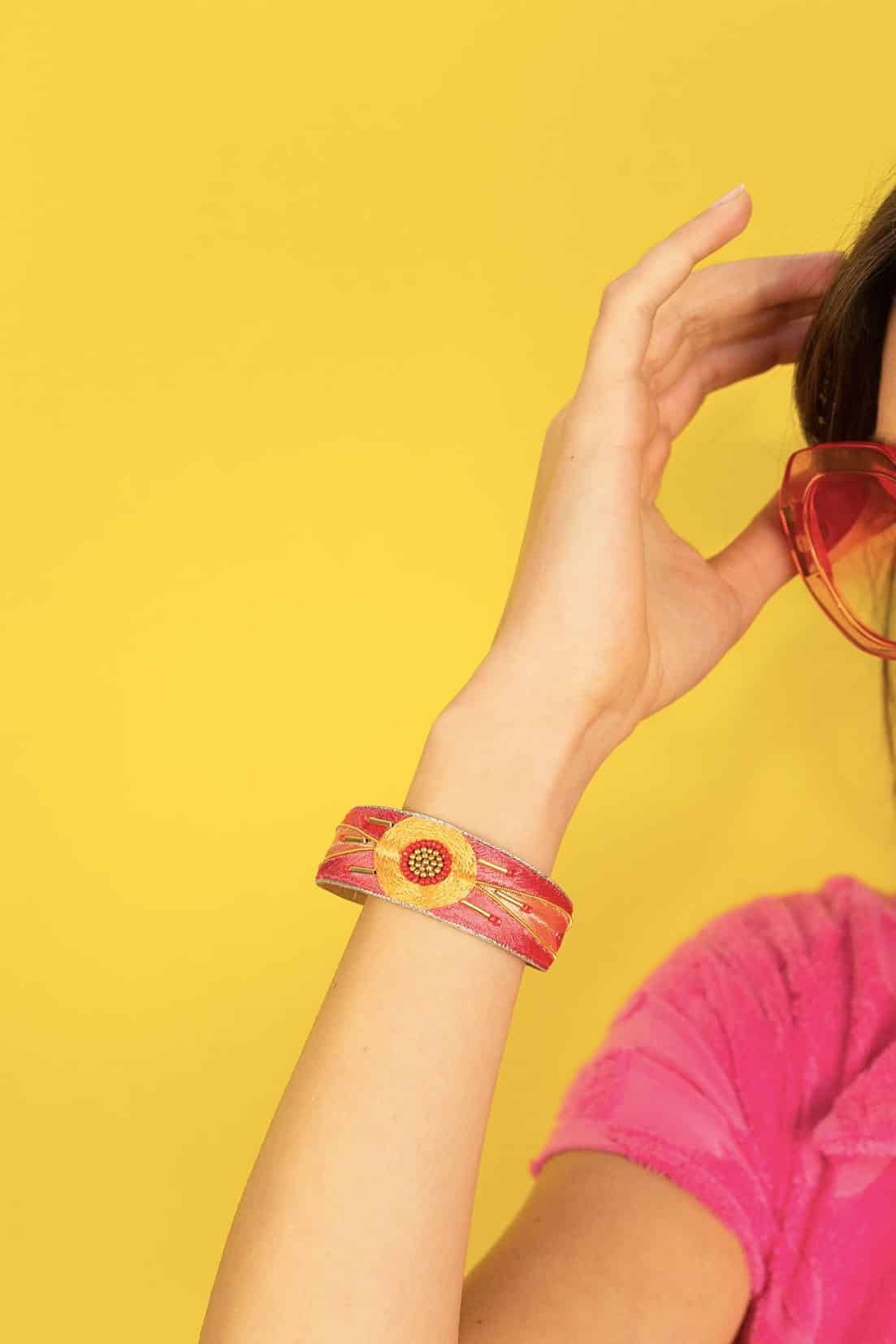 Femme dans un fond jaune, la main relevée vers la tête, portant un bangle fin ethnique brodé à la main de fil de soie couleur rose et orange. Perles en laiton, perles japonaises rouges, doublure en cuir souple.