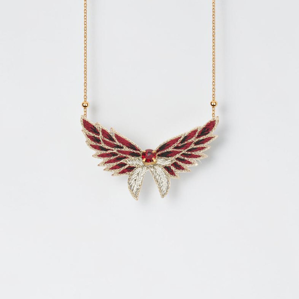 Médaillon Glanalia de la collection Glamrock en coloris Burgundy, pendentif brodé en forme d'ailes bordeaux et dorées avec cristal rouge, vue de face.